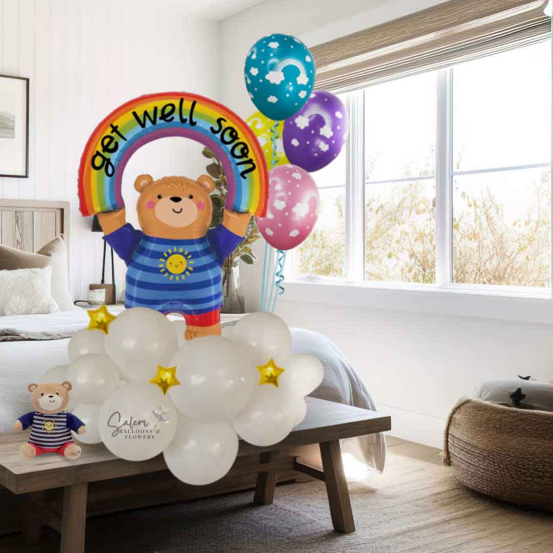 A charming teddy bear balloon holding a rainbow with a heartfelt "Get-Well-Soon" message stands on a cloud adorned with stars, dressed in a striped T-shirt and basking in the warmth of a bright sun standing on a bench in front of a bed in a bedroom. As a finishing touch, a cluster of vibrant rainbow balloons floats alongside.